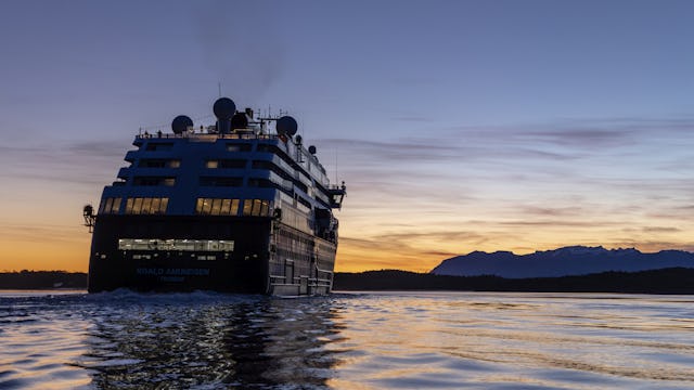 Läs mer om HX Hurtigruten Expeditions. Bilden visar ett av HX fartyg bakifrån i skymningen på vattnet.