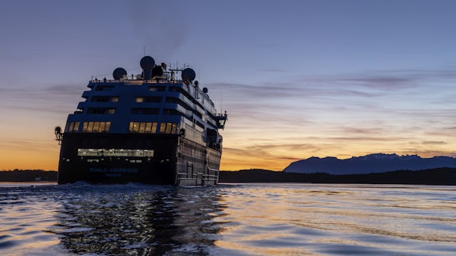 Läs mer om HX Hurtigruten Expeditions. Bilden visar ett av HX fartyg bakifrån i skymningen på vattnet.