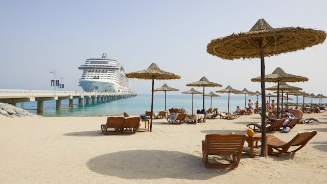 Bilden visar MSC Virtuosa som ligger i hamn vid en strand. Bilden har lite solstolar i förgrunden.