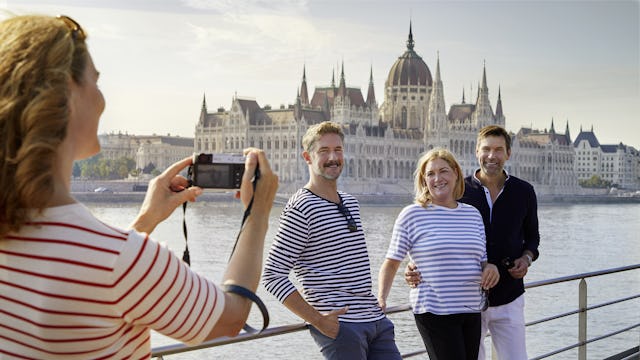 Bilden visar en person i förgrunden som tar en gruppbild på tre människor ombord Viva Tiara i Budapest med Ungerns parlamentbyggnad i bakgrunden. Kom nära och upptäck Europa på en flodkryssning.