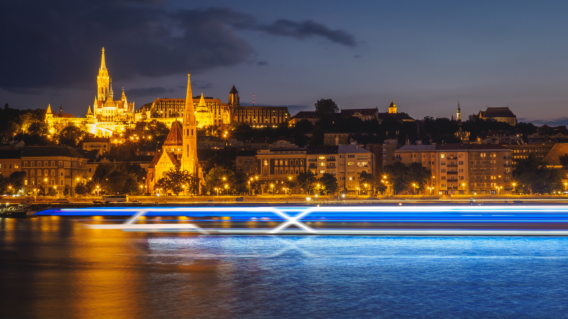 Celebrity Cruises börjar med flodkryssningar. Bilden visar Budapest i bakgrunden med ett suddigt flodkryssningsfartyg i förgrunden.