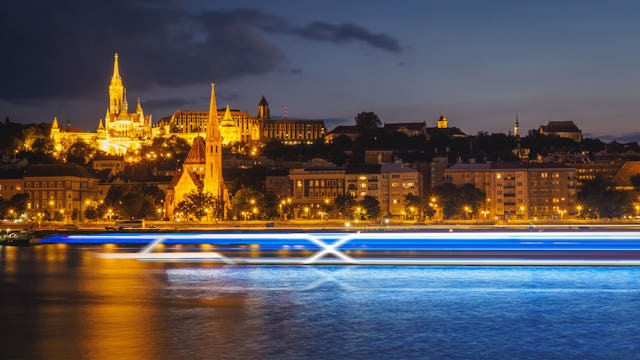 Celebrity Cruises börjar med flodkryssningar. Bilden visar Budapest i bakgrunden med ett suddigt flodkryssningsfartyg i förgrunden.