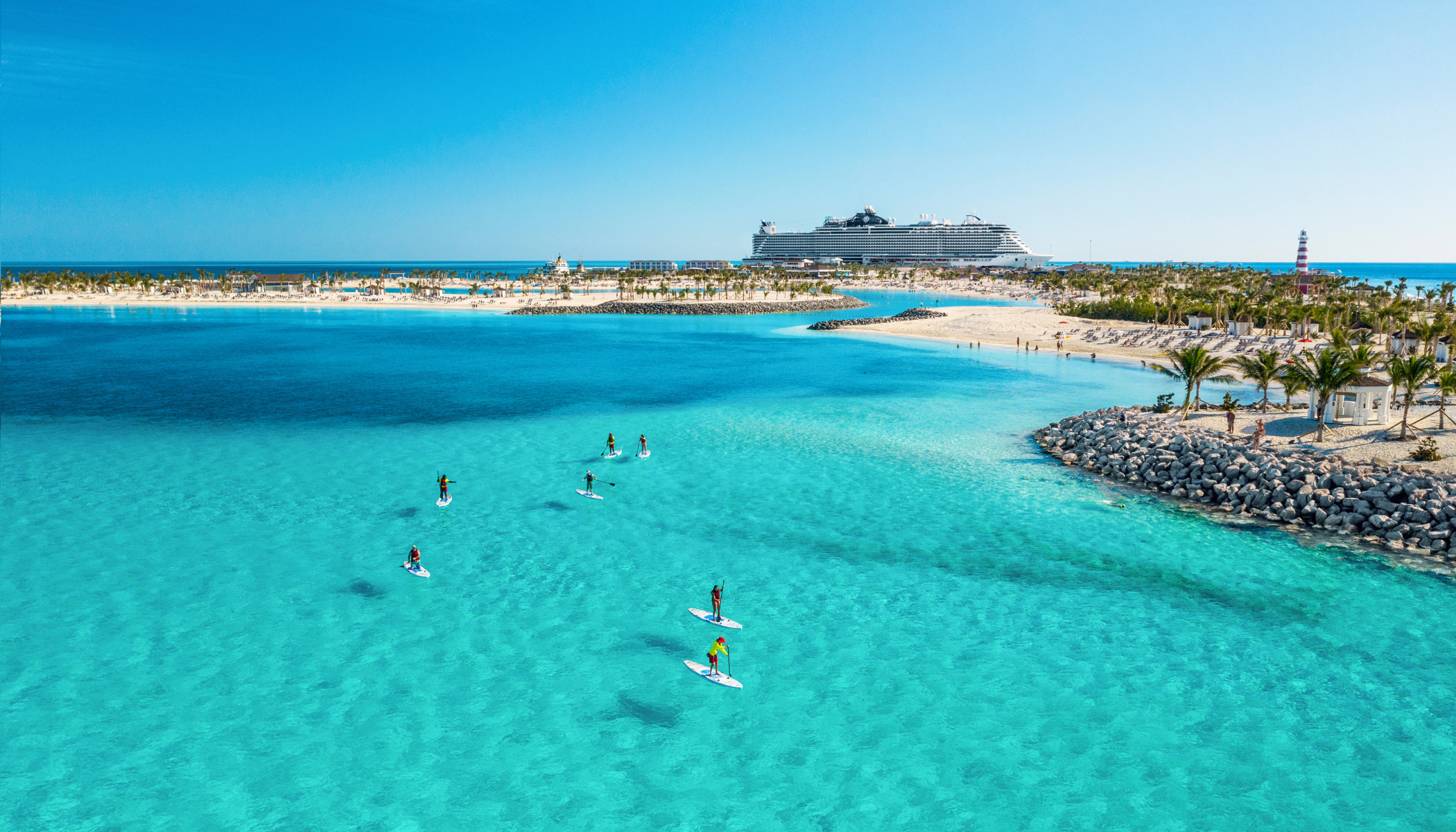 Boka din nästa kryssning hos Cruise Market. Bilden visar ett fartyg som ligger i hamn i Karibien. Man ser turkost vatten i förgrunden med folk som stand-up paddlar.