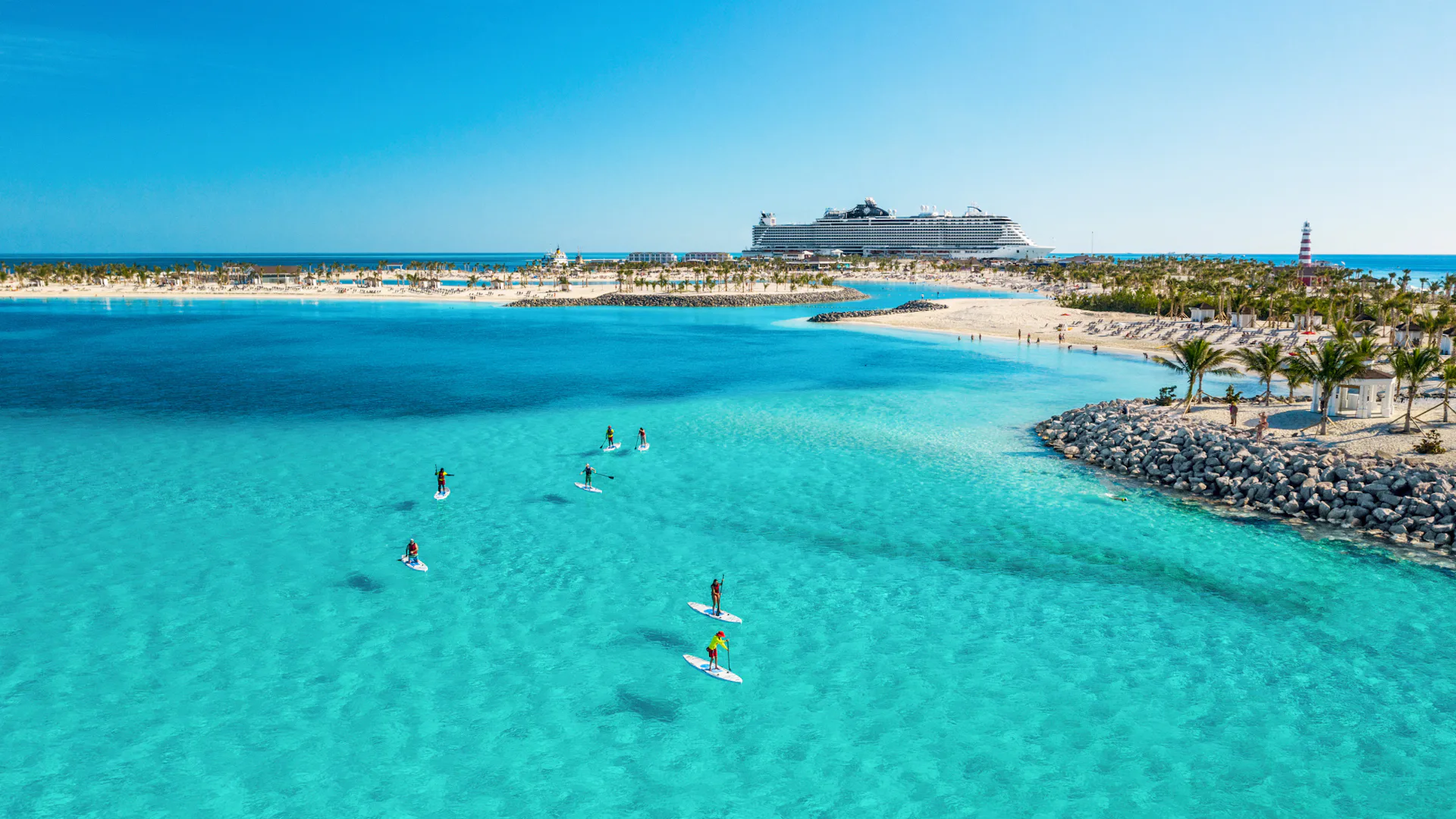 Boka din nästa kryssning hos Cruise Market. Bilden visar ett fartyg som ligger i hamn i Karibien. Man ser turkost vatten i förgrunden med folk som stand-up paddlar.