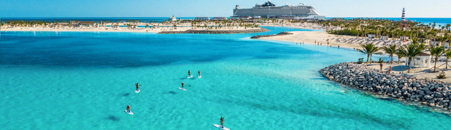 Boka din nästa kryssning hos Cruise Market. Bilden visar ett fartyg som ligger i hamn i Karibien. Man ser turkost vatten i förgrunden med folk som stand-up paddlar.