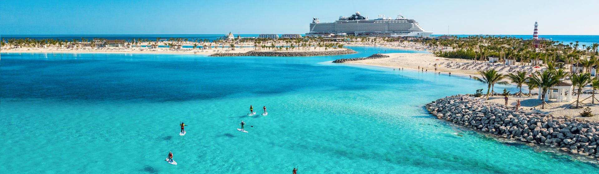 Boka din nästa kryssning hos Cruise Market. Bilden visar ett fartyg som ligger i hamn i Karibien. Man ser turkost vatten i förgrunden med folk som stand-up paddlar.