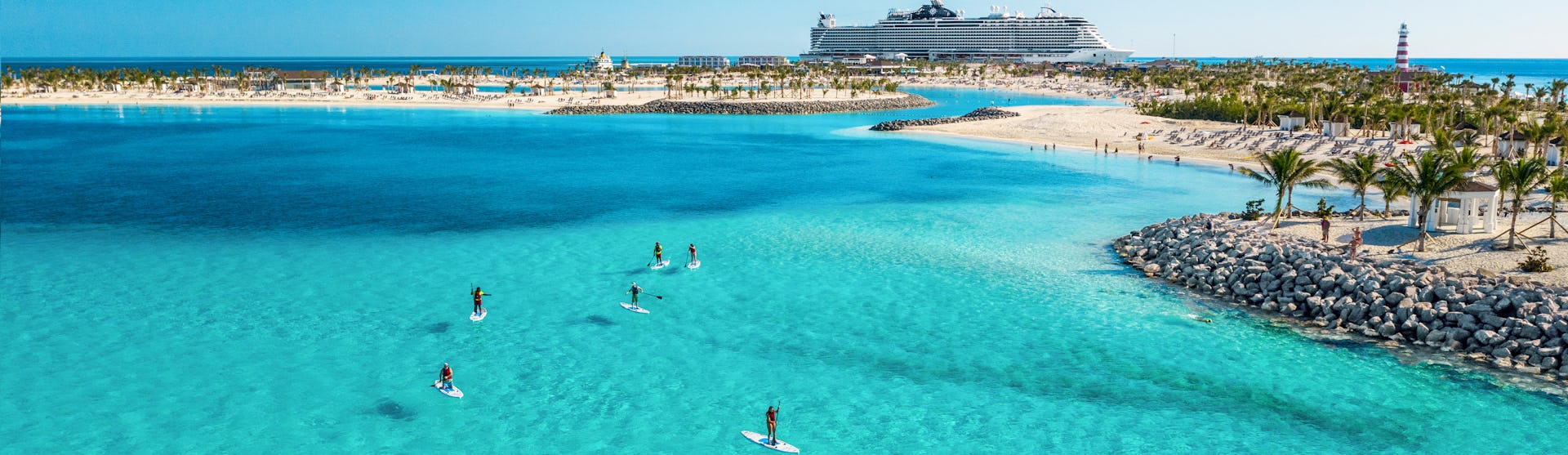 Boka din nästa kryssning hos Cruise Market. Bilden visar ett fartyg som ligger i hamn i Karibien. Man ser turkost vatten i förgrunden med folk som stand-up paddlar.