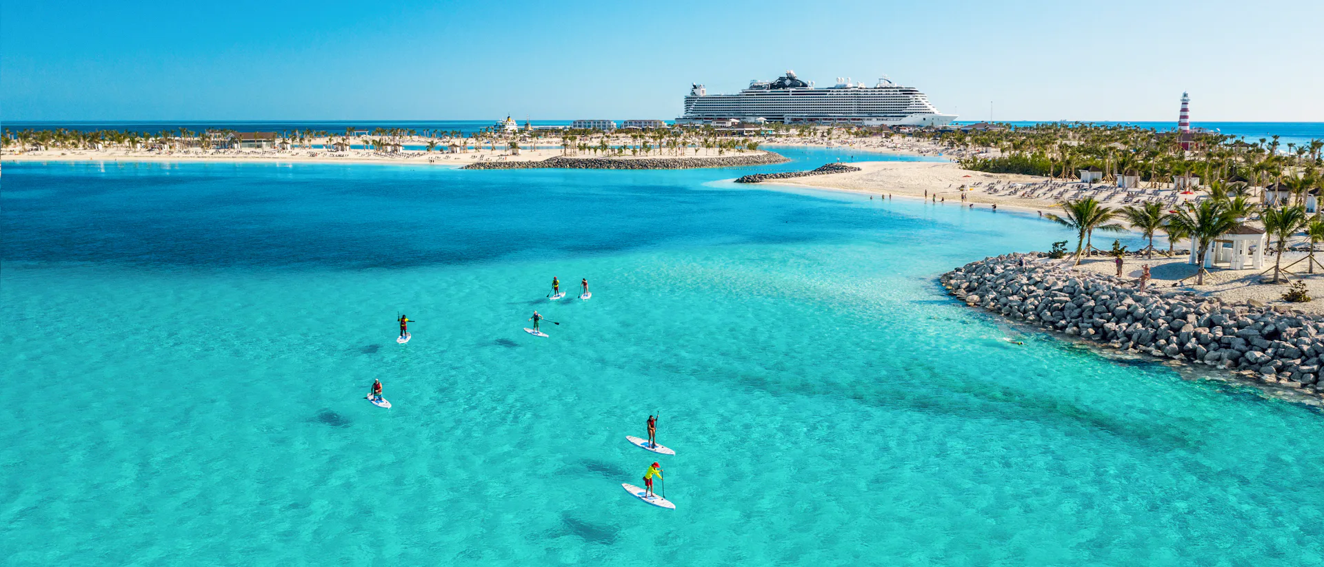Boka din nästa kryssning hos Cruise Market. Bilden visar ett fartyg som ligger i hamn i Karibien. Man ser turkost vatten i förgrunden med folk som stand-up paddlar.